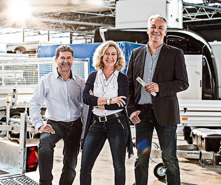 Een vrouw en twee mannen staan voor een selectie van verschillende Humbaur-trailers in de fabriekshal. | © Humbaur GmbH